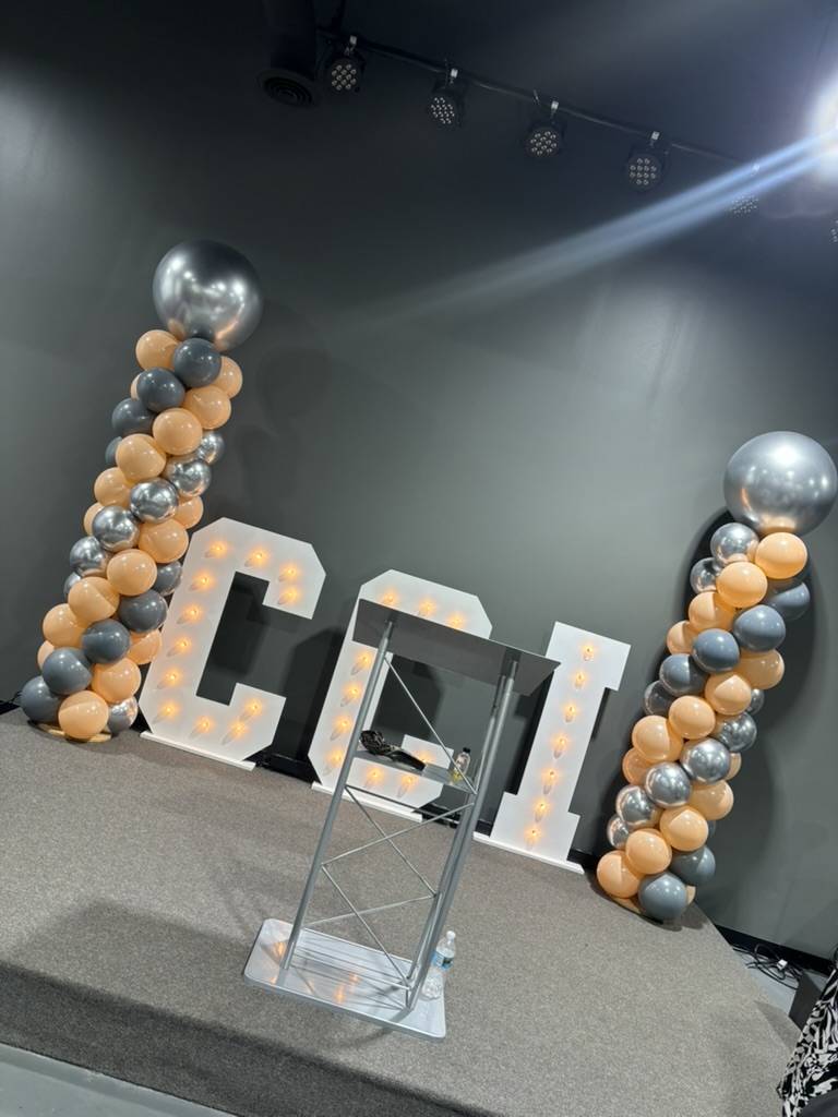 Corporate stage with lit marquee initials, balloon columns in the company's colors and a clear podium, styled by Sharp Designs